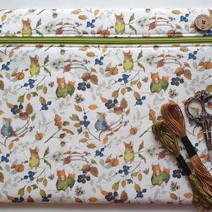 May include: A white zippered pouch with a whimsical pattern of mice, berries, and foliage. The pouch has a green zipper and a wooden button. Embroidery floss and decorative scissors are next to the pouch.