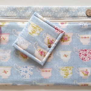 May include: A rectangular fabric pouch with a zipper closure and a smaller matching folded fabric wallet with a snap button. Both items feature a light blue background with a repeating pattern of various floral teacups and a floral top border.