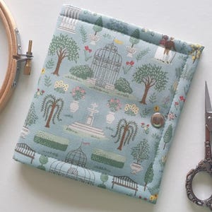 Può includere: Un astuccio per lavori di cucito trapuntato azzurro con una stampa a scena di giardino. L'astuccio ha una chiusura a scatto ed è accanto a un telaio da ricamo in legno e forbici decorative.