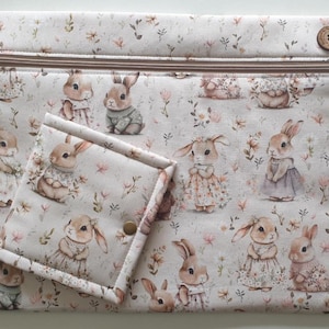 May include: A cream-colored zippered pouch and matching small wallet featuring a repeating pattern of adorable bunnies in dresses and sweaters, surrounded by floral accents. The pouch has a wooden button closure and a brown zipper.