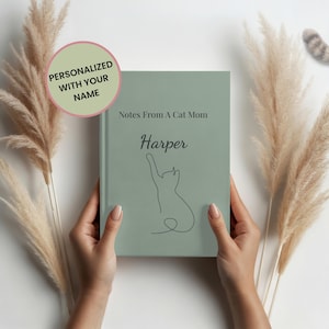 May include: A sage green notebook with the text "Notes From A Cat Mom" and the name "Harper". A line drawing of a cat is on the cover. The notebook is held in front of a white background with decorative dried plants.