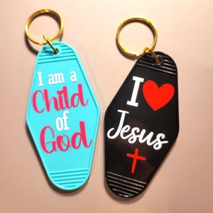 May include: Two keychains with gold rings. One is turquoise with the text "I am a Child of God" in pink and white. The other is black with "I ❤️ Jesus" in white, a red heart, and a red cross.
