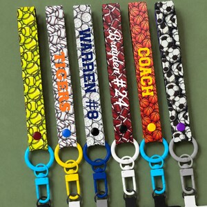 May include: Six fabric wristlet keychains featuring sports ball patterns including softball, baseball, volleyball, football, basketball, and soccer. Each strap displays custom text like TIGERS, WARREN #8, Brayden #24, and COACH with metal clasps.