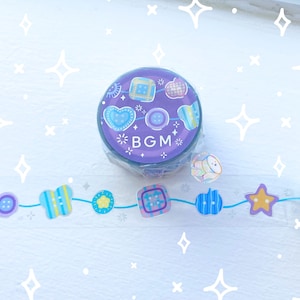 May include: A roll of purple washi tape with a design of hearts, squares, and stars, with the text "BGM". A strip of clear tape with matching designs is also visible. Decorative craft supplies.