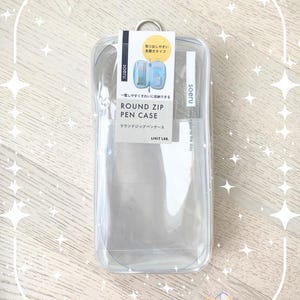 Può includere: Astuccio rettangolare trasparente con chiusura a zip e anello in metallo argentato. L'etichetta presenta testo giapponese, tra cui "ROUND ZIP PEN CASE". Progettato per una facile conservazione.