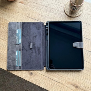 May include: Gray tablet case with a magnetic closure, open to reveal a tablet and card slots. The case is made of a textured material and is open on a wooden surface. The tablet screen is black.