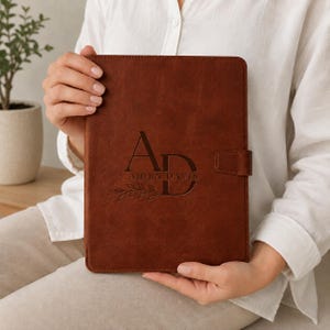May include: A rectangular brown leather portfolio with a magnetic side closure. The front cover features a debossed design with the large letters AD, the name AIDEN DAVIS, and a small leafy branch graphic centered on the smooth surface.