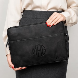 May include: A black rectangular laptop sleeve featuring a zippered closure and a front pocket. The surface is embossed with a circular monogram containing the letters MRG in the center and the name Emily printed in the bottom right corner.