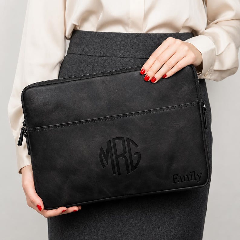 May include: A black rectangular laptop sleeve featuring a zippered closure and a front pocket. The surface is embossed with a circular monogram containing the letters MRG in the center and the name Emily printed in the bottom right corner.