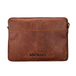 May include: A rectangular brown leather laptop sleeve with a smooth, matte finish and visible stitching along the top edge. The front features a horizontal leather patch embossed with the name MICHAEL in a bold, dark, sans-serif font.