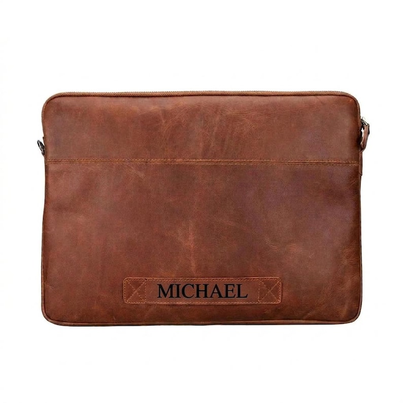 May include: A rectangular brown leather laptop sleeve with a smooth, matte finish and visible stitching along the top edge. The front features a horizontal leather patch embossed with the name MICHAEL in a bold, dark, sans-serif font.