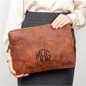 May include: A rectangular brown leather laptop sleeve featuring a zippered closure and a front pocket. The center of the sleeve is embossed with a circular monogram containing the letters MRG, and the name Emily is engraved in the bottom right corner.