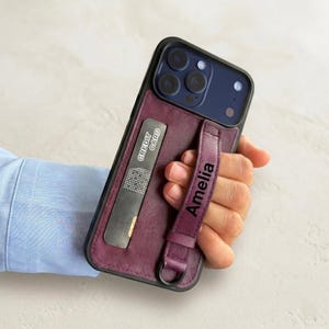 May include: A dark purple phone case with a black border, featuring a card slot and a strap with the name "Amelia" printed on it. The case also has a "CREDIT CARD" label.