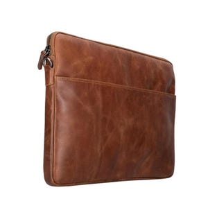 May include: A rectangular brown leather laptop sleeve featuring a smooth, textured surface with visible grain patterns. The sleeve includes a zippered closure along the top edge with a metal pull tab and a slim front pocket compartment for storage.