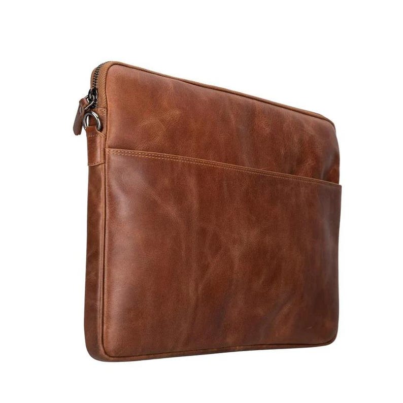 May include: A rectangular brown leather laptop sleeve featuring a smooth, textured surface with visible grain patterns. The sleeve includes a zippered closure along the top edge with a metal pull tab and a slim front pocket compartment for storage.