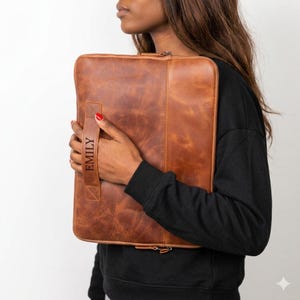 May include: A rectangular brown leather laptop sleeve featuring a vertical stitched seam and a side handle. The handle is embossed with the name EMILY in capital letters. The sleeve has a smooth, matte finish with visible natural grain texture.