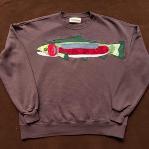 Può includere: Una felpa marrone con un'applicazione di pesce colorata. Il design del pesce include accenti verdi, rossi e blu. La felpa ha un girocollo e maniche lunghe. Il nome del marchio è visibile sul colletto.