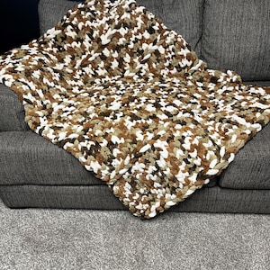 May include: A chunky knit blanket in shades of brown, white, and tan, draped over a gray sofa. The blanket's textured surface creates a cozy, inviting look. This handmade throw blanket is perfect for adding warmth and style to any living space.
