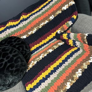 May include: A chunky knit blanket in a striped pattern of black, orange, yellow, brown, green, and white. The blanket is folded over a gray sofa, with a black patterned pillow visible.