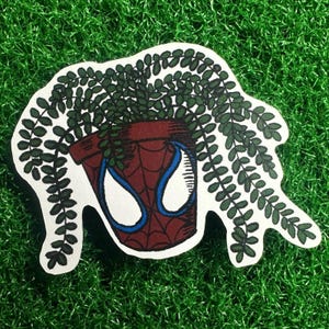 Puede incluir: Una pegatina que representa una maceta marrón con un diseño de máscara de Spider-Man, llena de plantas verdes. La pegatina tiene un borde blanco y está sobre un fondo verde y herboso.