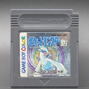 May include: A gray Game Boy Color cartridge with a silver label featuring a blue and white Pokemon character. The label reads "Pocket Monsters Silver" in Japanese and "Game Boy & Color Common" in English. The cartridge is for the Japanese market and has the code DMG-AAXJ-JPN.