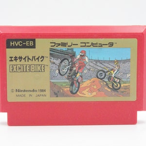 May include: A red Nintendo Entertainment System game cartridge for the game "Excitebike". The cartridge has a gold label with the game title in Japanese and English, along with the Nintendo logo and copyright information. The game features a motocross race theme with two riders on dirt bikes jumping over a ramp.