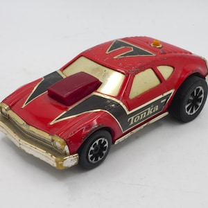 May include: A red Tonka toy car with black and gold accents. The car has a red hood scoop and a black stripe down the side. The car is in good condition with some minor wear and tear.