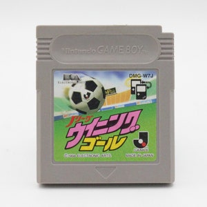 May include: A gray Game Boy Color cartridge with a green, pink, and white label. The label features a soccer ball, the text "J-League Soccer", and the text "C 1994 ELECTRONIC ARTS MADE IN JAPAN".