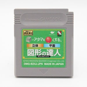 May include: A gray Nintendo Game Boy cartridge with a green and gold label. The label features Japanese text and illustrations of a cube and a red sphere. The cartridge is labeled "図形の達人" and "MADE IN JAPAN."