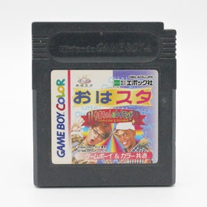 May include: A black Game Boy Color cartridge with a colorful label featuring the text "Oha-Sta" and images of two people. The label also includes the text "Game Boy & Color Common" in Japanese.
