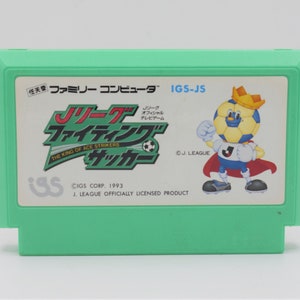 May include: A mint green video game cartridge featuring the game "J. League Fighting Soccer". The label includes Japanese text, a soccer ball mascot wearing a crown and cape, and the text "THE KING OF ACE STRIKERS".
