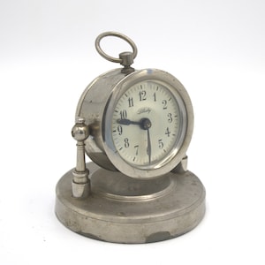 May include: A small, silver-toned desk clock with a round face and a circular handle on top. The clock face is white with black numerals and hands. The base is circular and the clock is supported by two decorative posts.