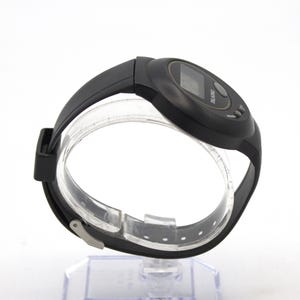 May include: Black digital talking watch with a black strap. The watch features a digital display and two buttons. The word "TALKING" is printed on the watch face. The watch is displayed on a clear plastic stand.