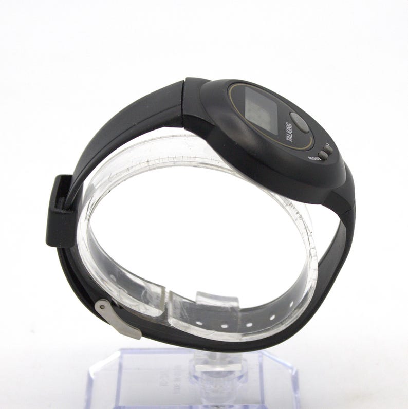 May include: Black digital talking watch with a black strap. The watch features a digital display and two buttons. The word "TALKING" is printed on the watch face. The watch is displayed on a clear plastic stand.