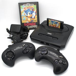 May include: A black Sega Genesis console with two controllers, a game cartridge, and a game box featuring Sonic the Hedgehog. The game cartridge and box both have the text "NOT FOR RESALE".