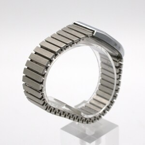 May include: A silver-toned wristwatch featuring a flexible, expandable metal link band with a brushed finish. The watch case is rectangular with rounded edges and a smooth, metallic surface, showcasing a classic design with a continuous link structure.