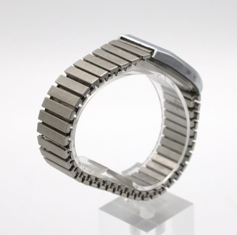 May include: A silver-toned wristwatch featuring a flexible, expandable metal link band with a brushed finish. The watch case is rectangular with rounded edges and a smooth, metallic surface, showcasing a classic design with a continuous link structure.