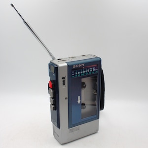 May include: A vintage Sony FM/AM radio cassette player with a silver and blue design. The portable device features a retractable antenna, a cassette tape window, and control buttons. The radio dial is visible, and the brand name "SONY" is printed on the top.