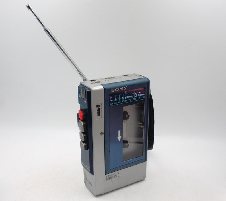 May include: A vintage Sony FM/AM radio cassette player with a silver and blue design. The portable device features a retractable antenna, a cassette tape window, and control buttons. The radio dial is visible, and the brand name "SONY" is printed on the top.