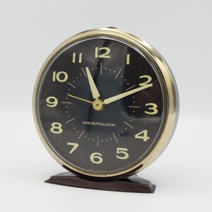 May include: A vintage Westclox alarm clock with a black face and gold-colored hands and numbers. The clock has a brown plastic base.