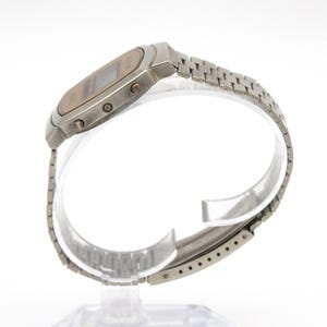 May include: A vintage digital wristwatch with a silver-tone metal band and case. The watch face is a light brown color with a digital display. The band is made of small, connected metal links. The watch is displayed on a clear stand.