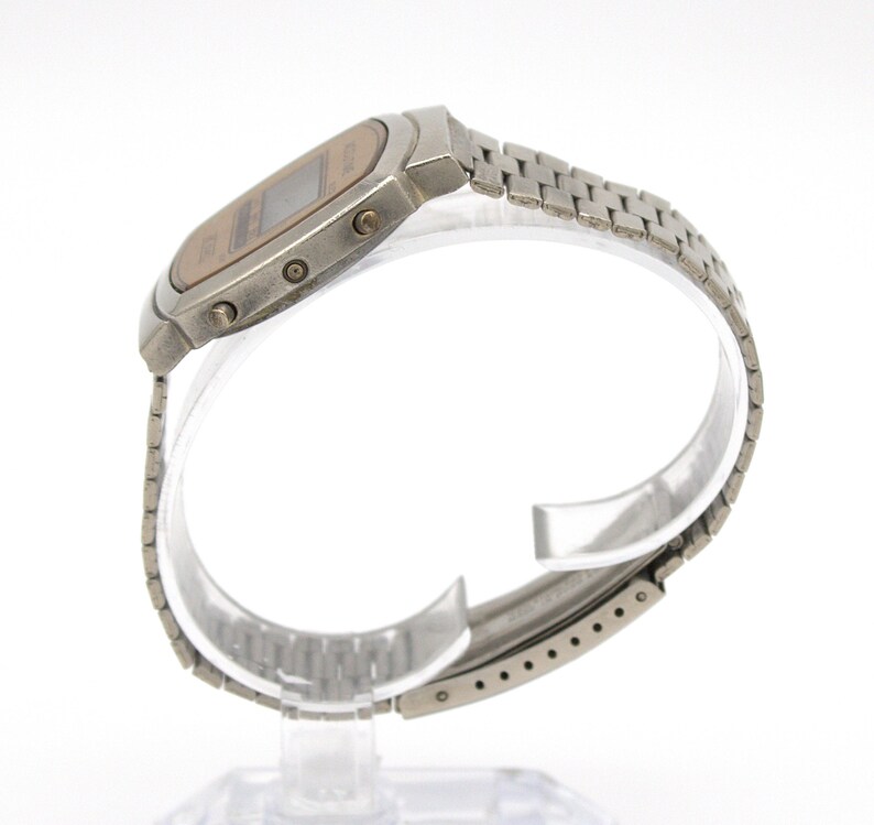 May include: A vintage digital wristwatch with a silver-tone metal band and case. The watch face is a light brown color with a digital display. The band is made of small, connected metal links. The watch is displayed on a clear stand.