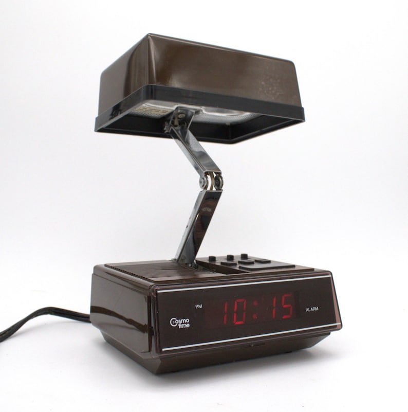 May include: A brown Cosmo Time digital alarm clock with a rectangular base and a hinged, adjustable lamp arm extending upward to a dark brown rectangular light shade. The front display shows the red digital time 10:15 with a PM indicator.