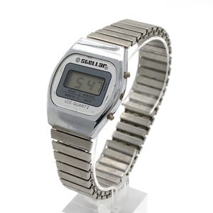 May include: A silver-toned digital wristwatch with a metallic expansion band. The watch face features the text STELLAR at the top and LCD QUARTZ at the bottom, with a digital display showing 5:47 and labels for hour, month, min/sec, and date.