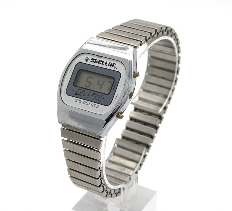 May include: A silver-toned digital wristwatch with a metallic expansion band. The watch face features the text STELLAR at the top and LCD QUARTZ at the bottom, with a digital display showing 5:47 and labels for hour, month, min/sec, and date.