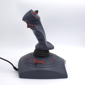 Puede incluir: Un joystick gris con botones y detalles en rojo. El joystick tiene un dise&ntilde;o alto y ergon&oacute;mico con una base negra y un cable negro que se extiende desde el lateral. Los botones est&aacute;n ubicados en la parte superior e inferior del joystick.