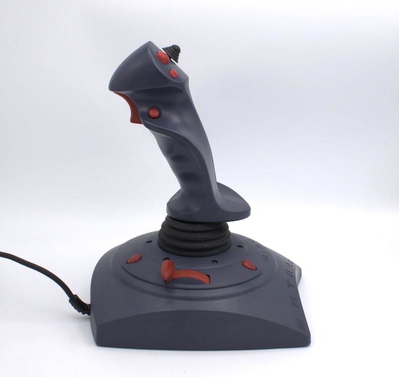 Puede incluir: Un joystick gris con botones y detalles en rojo. El joystick tiene un dise&ntilde;o alto y ergon&oacute;mico con una base negra y un cable negro que se extiende desde el lateral. Los botones est&aacute;n ubicados en la parte superior e inferior del joystick.