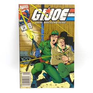 May include: A vintage G.I. Joe comic book cover featuring a dynamic action scene. The cover displays the title "G.I. Joe: A Real American Hero!" with a red star. The artwork depicts two figures in military attire, one firing a weapon. The comic is set against a brown background.