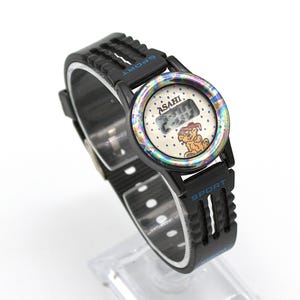 May include: Black digital watch with a black strap. The watch face has a white background with a cartoon dog and the word "ASAHI". The watch has the word "SPORT" in blue on the strap.
