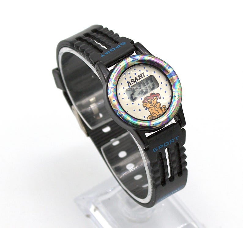 May include: Black digital watch with a black strap. The watch face has a white background with a cartoon dog and the word "ASAHI". The watch has the word "SPORT" in blue on the strap.
